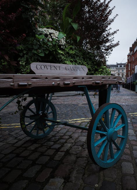 A vintage wooden cart with bright blue spoked wheels is positioned on a cobblestone street in Covent Garden. The cart serves as a decorative garden feature, with lush green foliage and white flowering