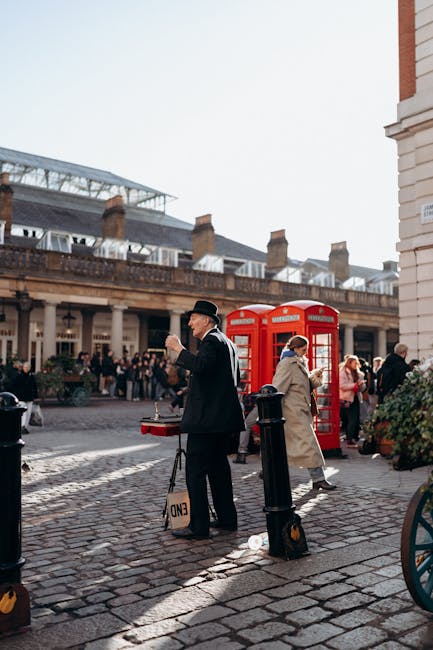 A vintage wooden cart with bright blue spoked wheels is positioned on a cobblestone street in Covent Garden. The cart serves as a decorative garden feature, with lush green foliage and white flowering